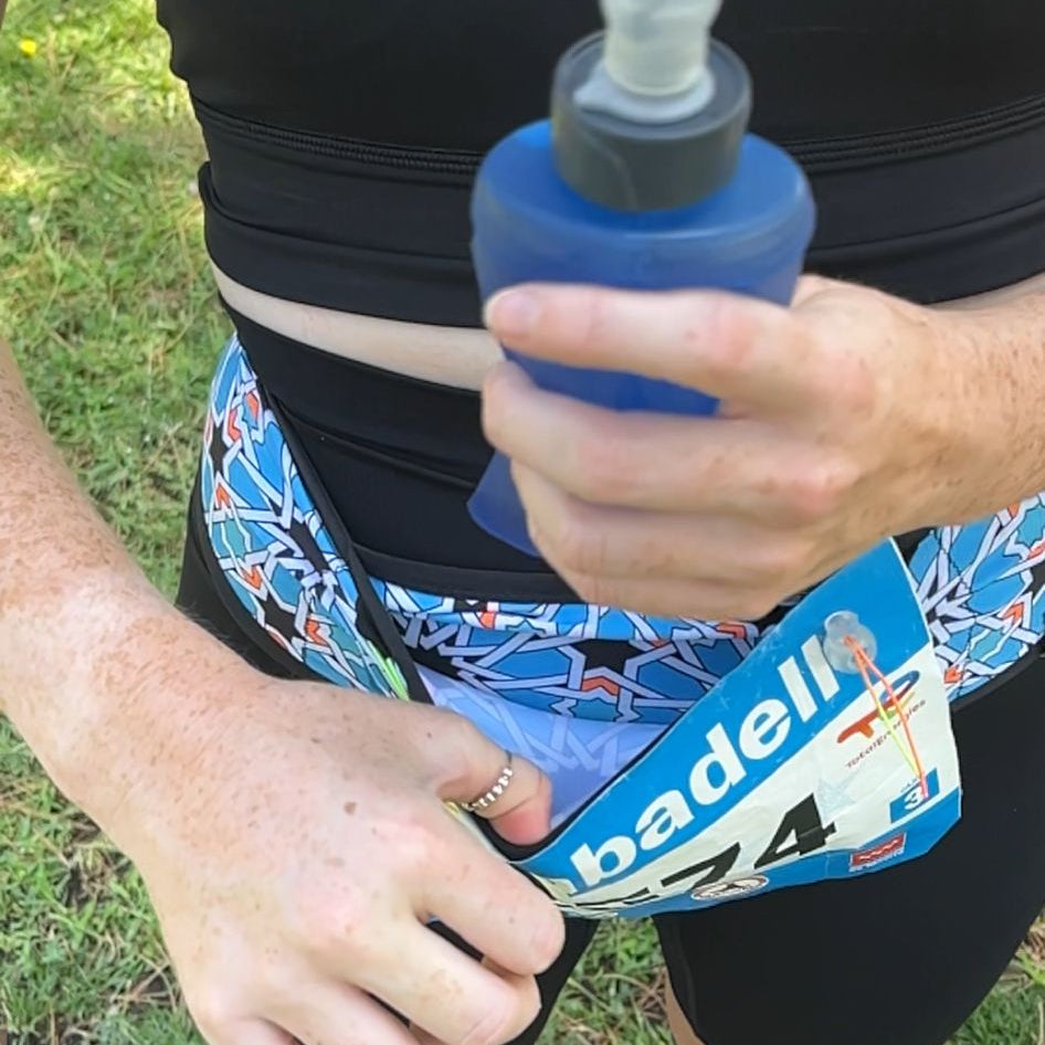 A person outdoors wearing a black athletic top and shorts, holding a blue spray bottle, with a blue and white running bib around their waist. Grass is visible in the background.