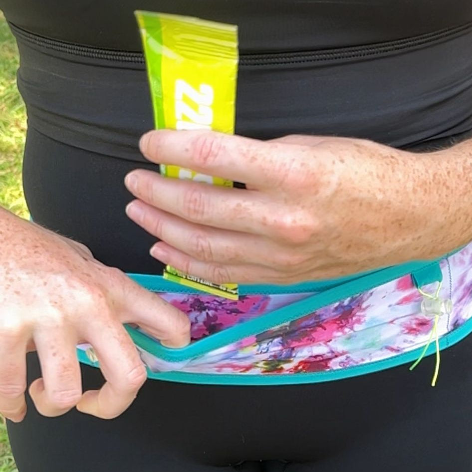 A person outdoors wearing a black athletic outfit and holding a bright yellow-green tube of Zinwa sunscreen over a colorful floral waist bag.