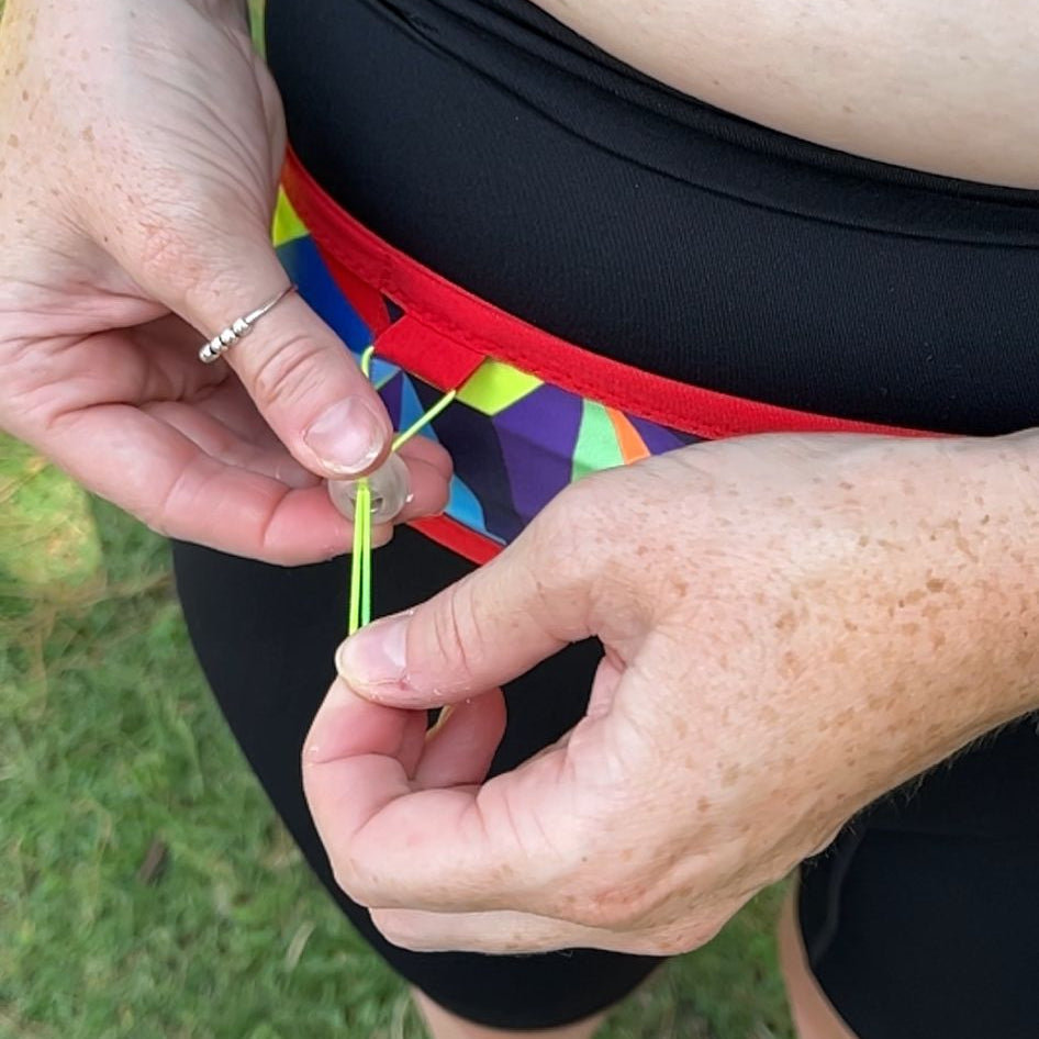A close-up lifestyle shot of a person outdoors wearing a black athletic waist wrap or belt with a colorful geometric pattern, adjusting a small green tag on the front.