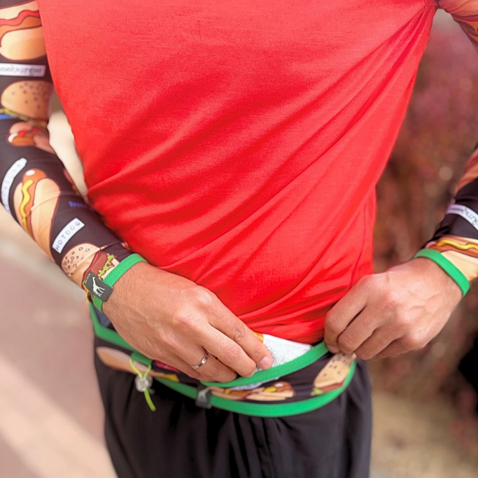 A close-up of a person wearing a red athletic shirt with a green waistband and a built-in pouch holding a white item, paired with black shorts and patterned long sleeves.