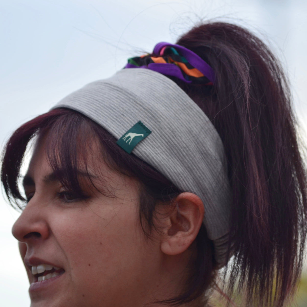 A close-up side profile of a woman wearing a light gray athletic headband with a small green logo patch, holding her hair in a ponytail with a purple and orange scrunchie, against a blurred outdoor background.