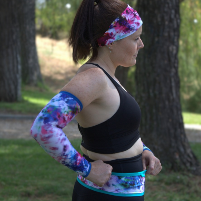 A woman in a park wearing a matching athletic set with a floral print. The set includes a long-sleeve arm sleeve, a waist band, a headband, and a sports bra, all featuring a pink, purple, and blue floral pattern with a bright turquoise blue trim.