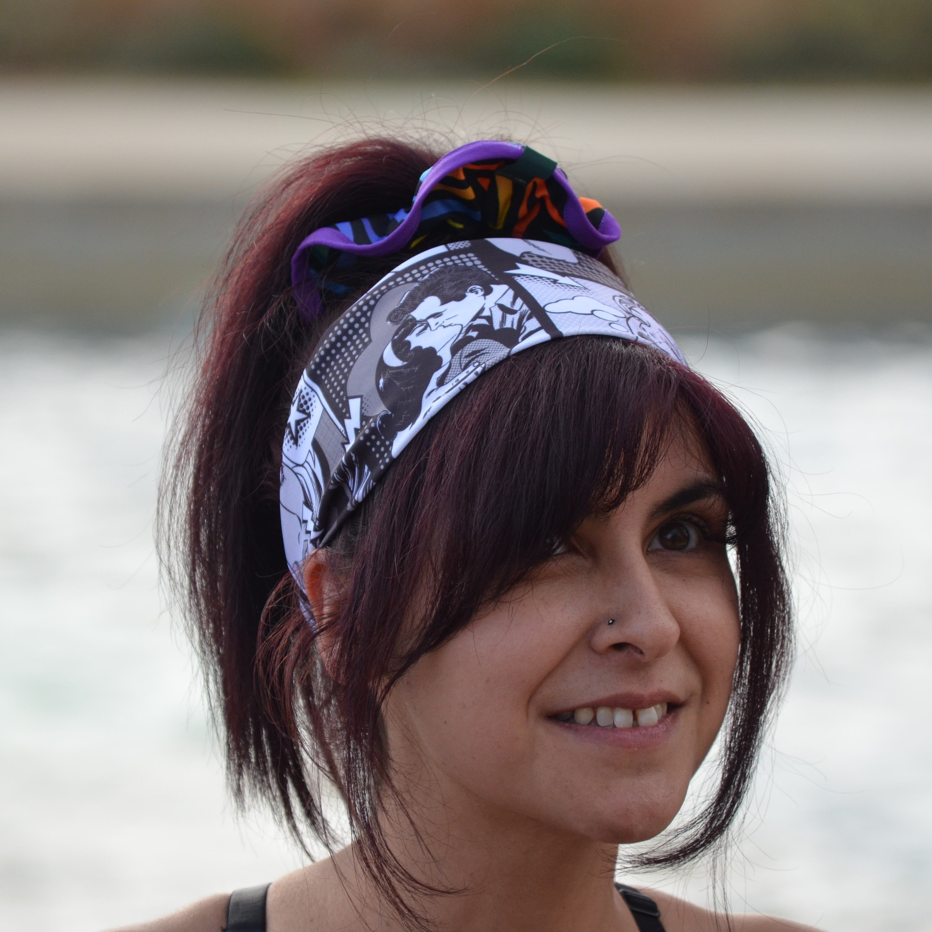 A woman with dark hair wearing a black and white patterned headband with a purple and orange scrunchie, photographed outdoors with a blurred water background.