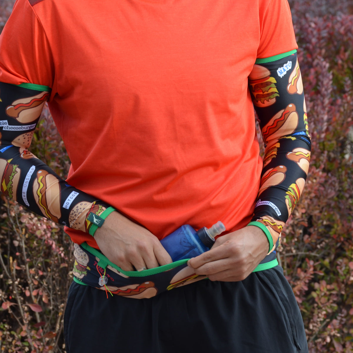 A close-up of a person outdoors wearing a bright orange athletic shirt with a green waist pocket, holding a blue water bottle. The shirt is layered over a black long-sleeve base layer with a colorful autumn leaf pattern in orange, brown, and green.