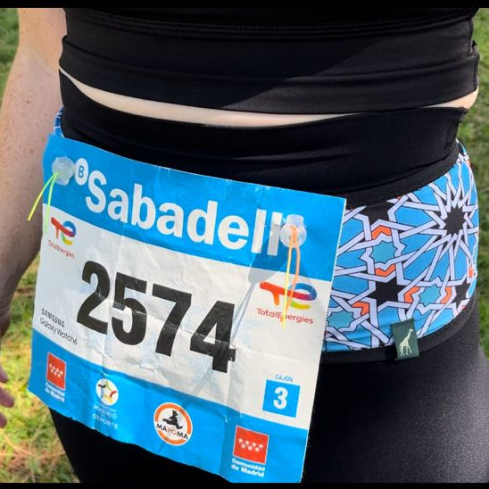 A close-up of a person outdoors wearing a blue and white Sabadel marathon bib number 2574 around the waist, with grass visible in the background.