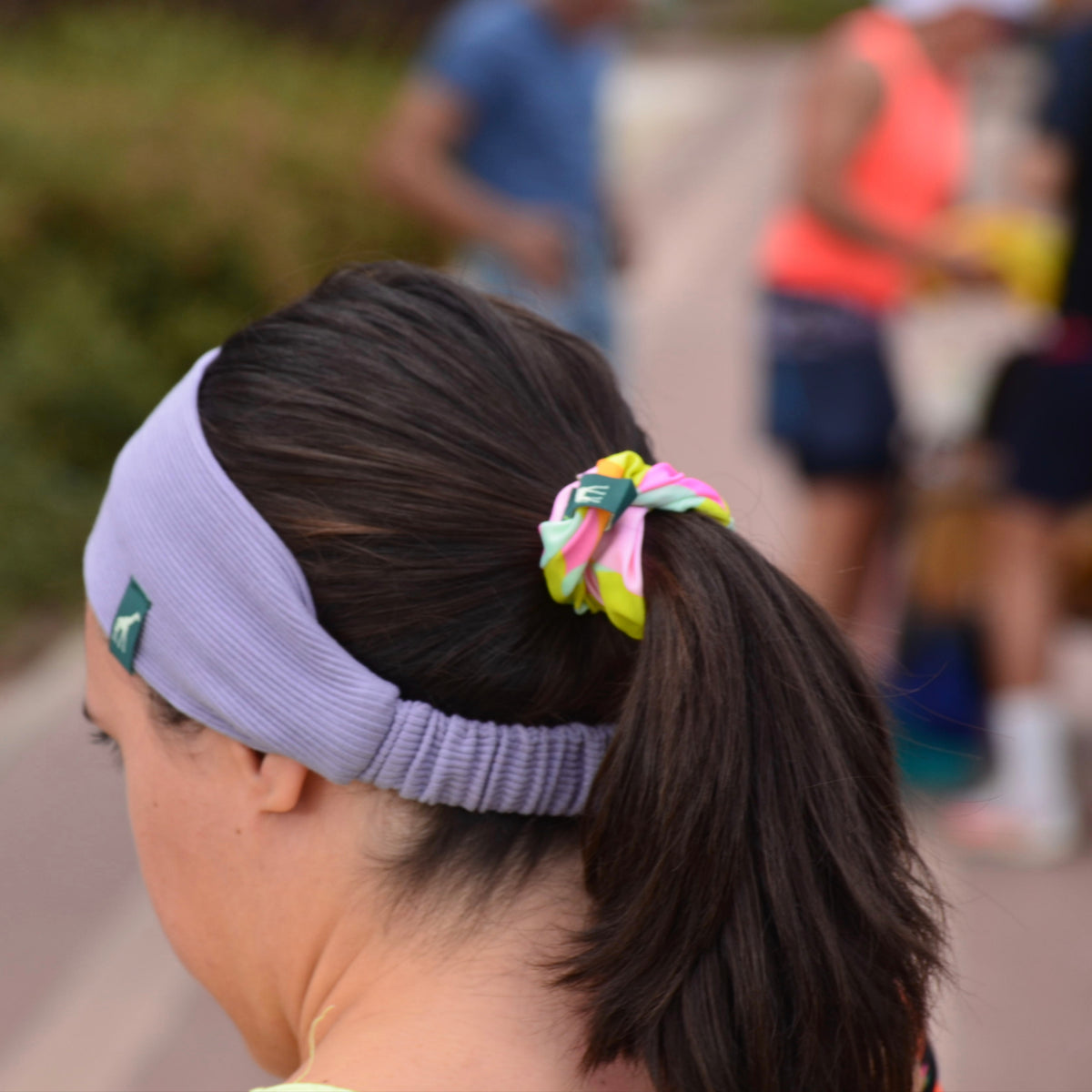 A woman outdoors wearing a light purple athletic headband with a small green logo and a colorful neon scrunchie holding her dark brown ponytail.
